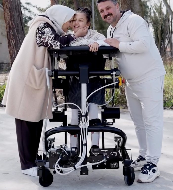 Yürüme robotunu kullanan bir çocuk, anne ve babasıyla birlikte açık havada