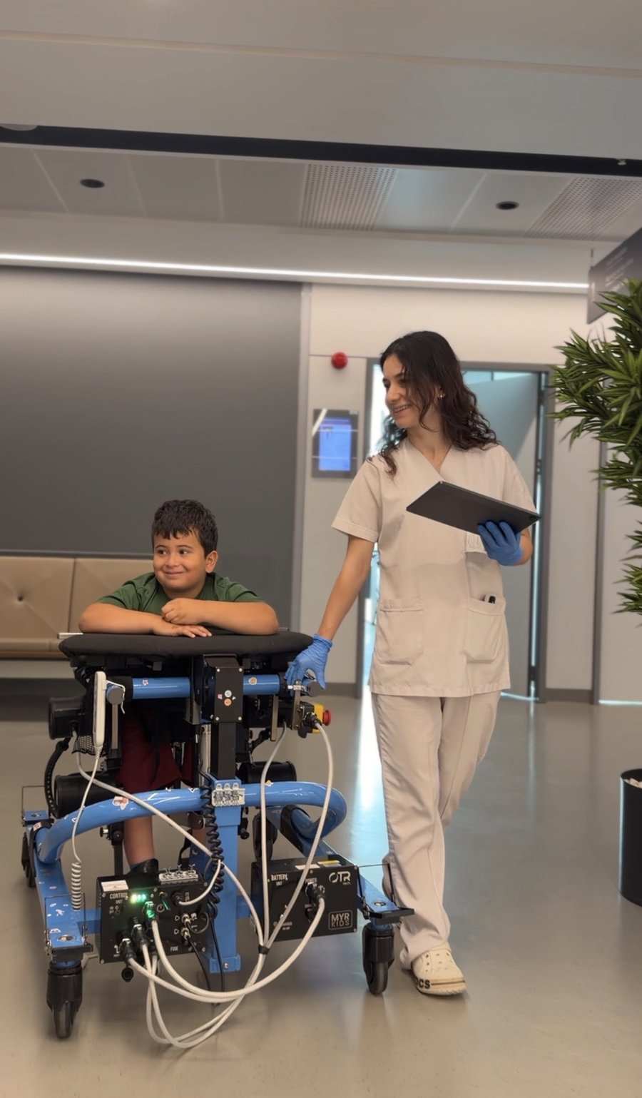 Klinik koridorunda yürüme robotunu kullanan bir çocuk ve yanında sağlık personeli